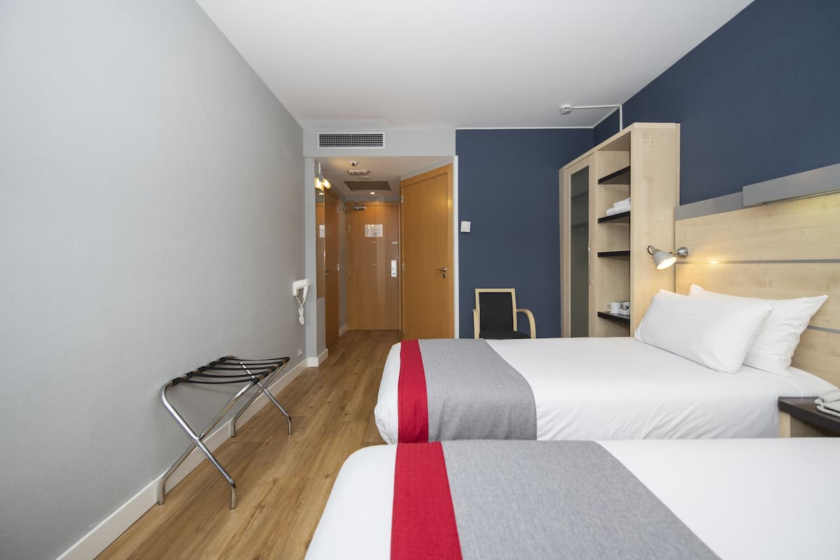 A hotel room with two beds, a wooden wardrobe, a small table, and a wall-mounted shelf.