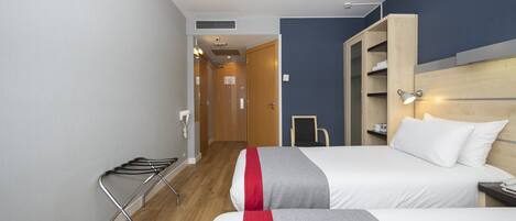 A hotel room with two beds, a wooden wardrobe, a small table, and a wall-mounted shelf.