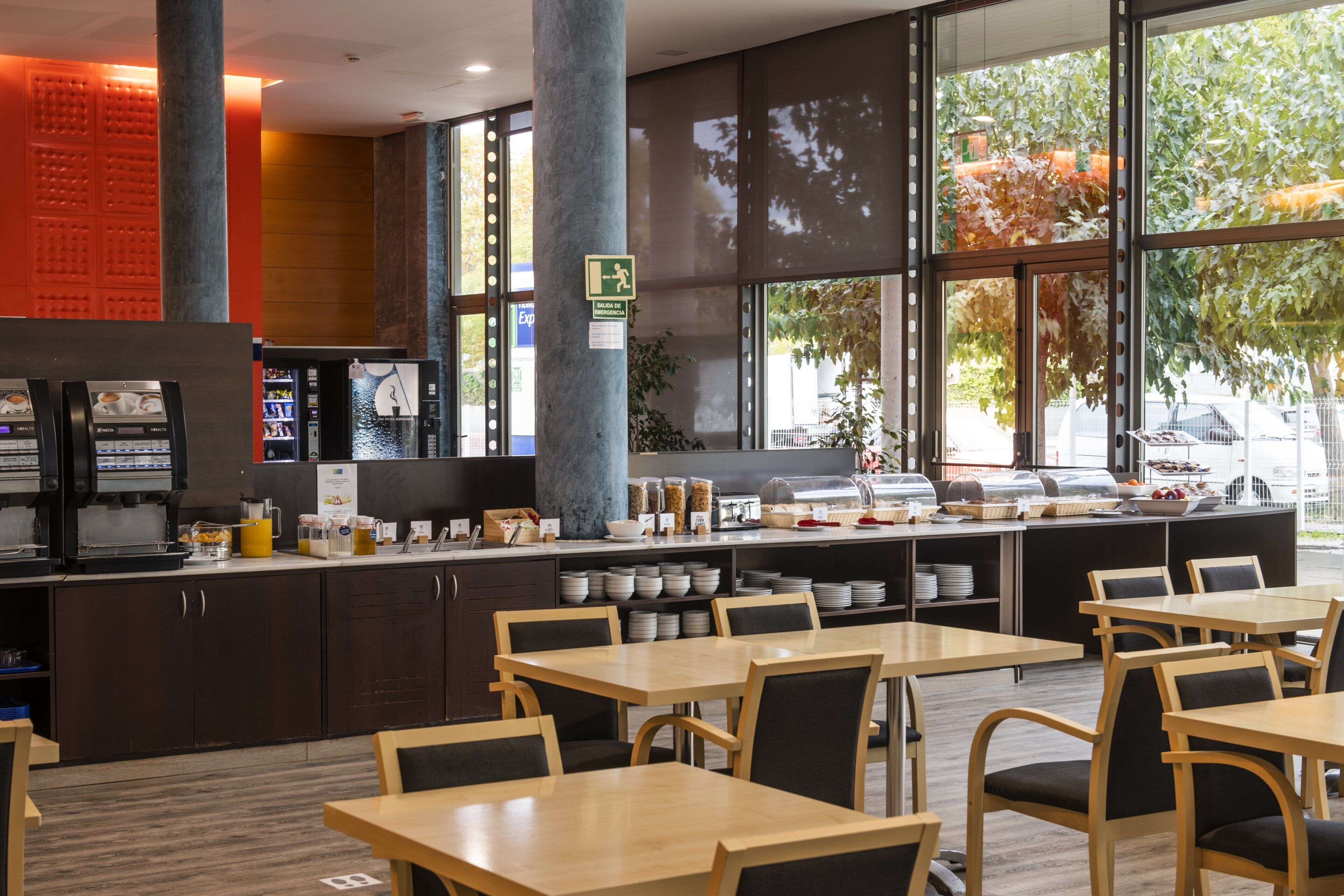Un café moderno con mesas y sillas de madera, una máquina de café y un mostrador de buffet con diversas opciones de comida.