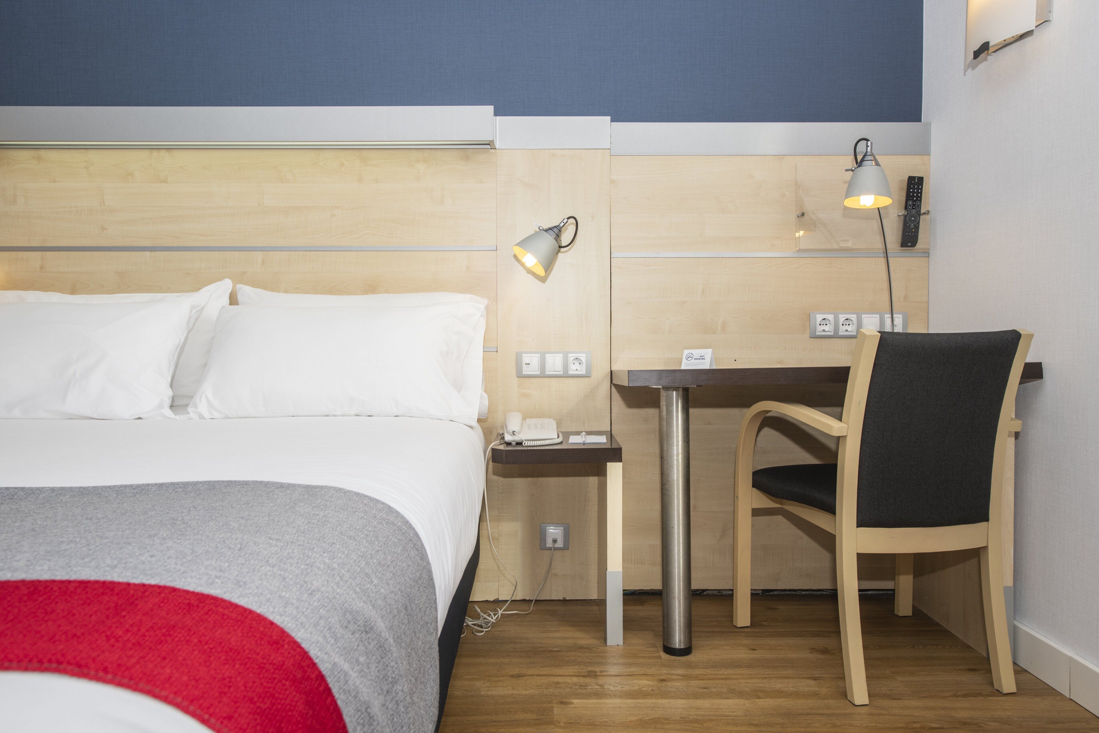 A hotel room with a bed, a desk, a chair, and a lamp.