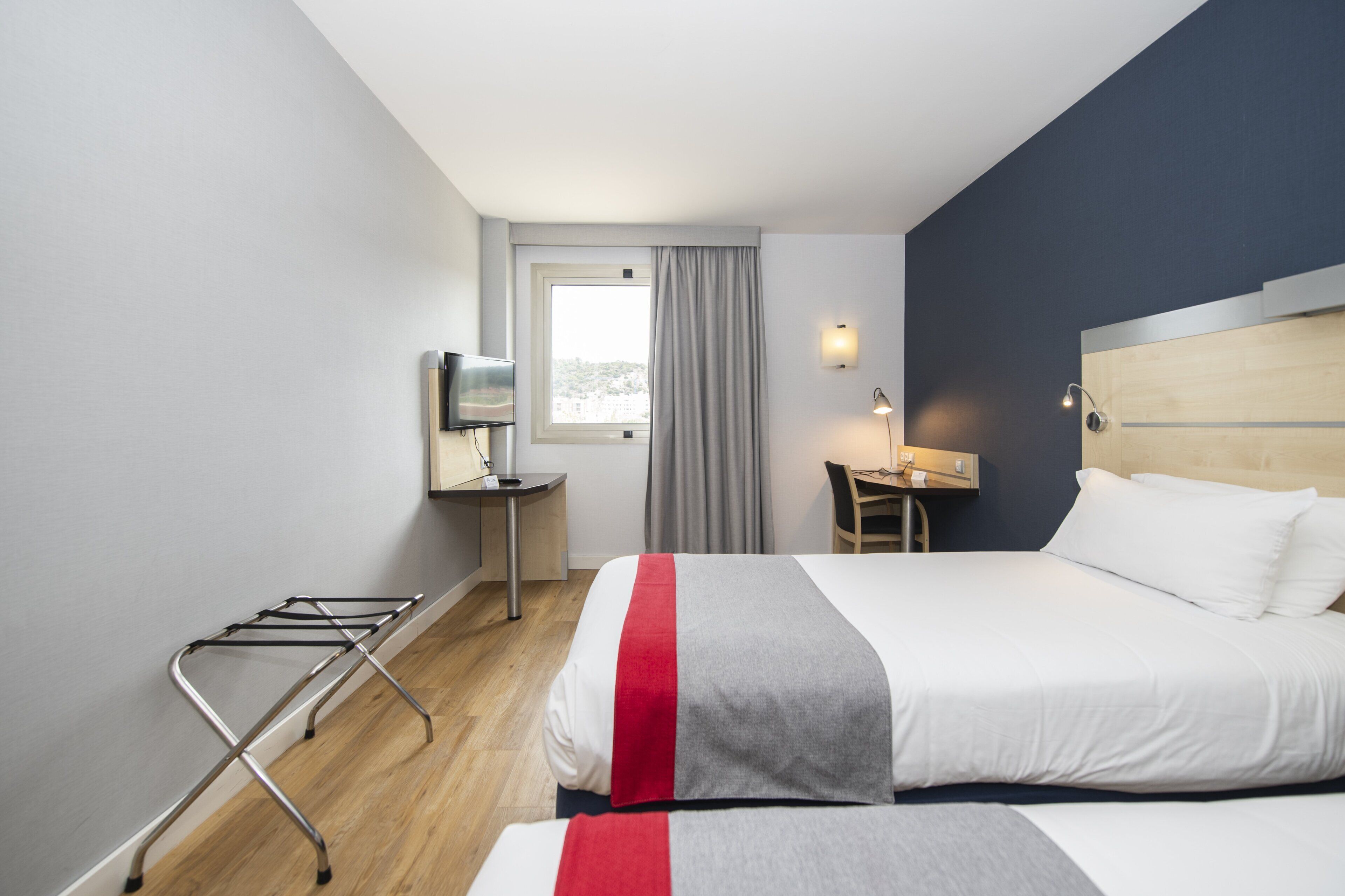 A hotel room with a bed, a desk, a chair, a TV, and a window with curtains.