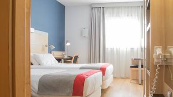 Et hotelværelse med to senge, et skrivebord, en stol, en lampe, en telefon og et vindue med gardiner.