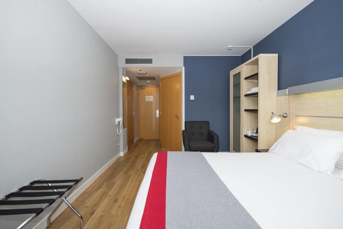 A hotel room with a bed, a chair, a wooden wardrobe, a desk, and a small table.