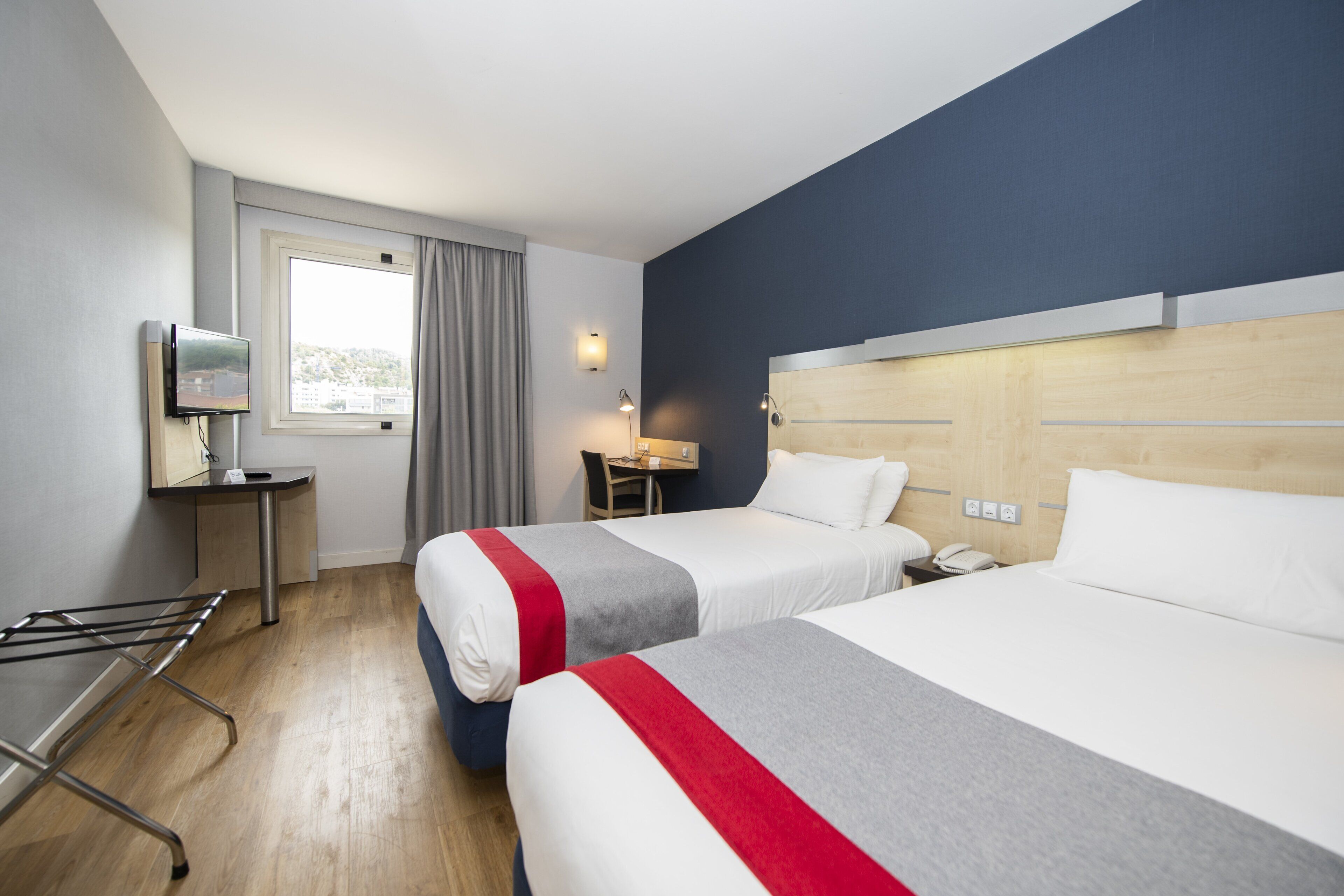 Ein Hotelzimmer mit zwei Betten, einem Schreibtisch, einem Stuhl, einem Fernseher und einem Fenster mit Vorhängen.