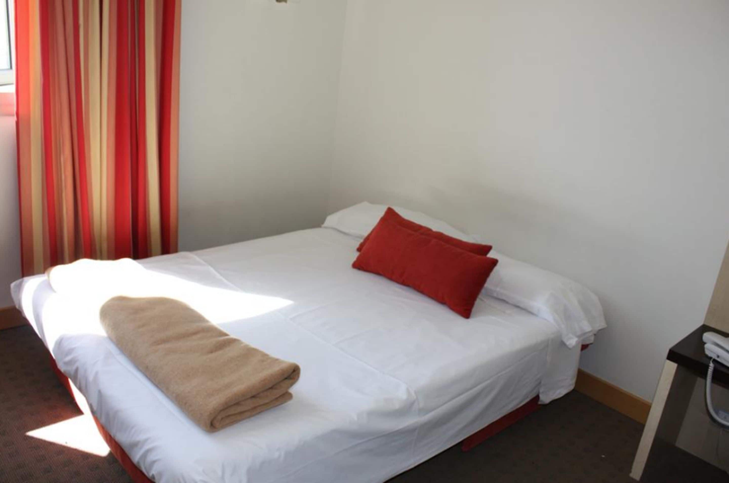 A single bed with white bedding, a red pillow, and a beige towel.