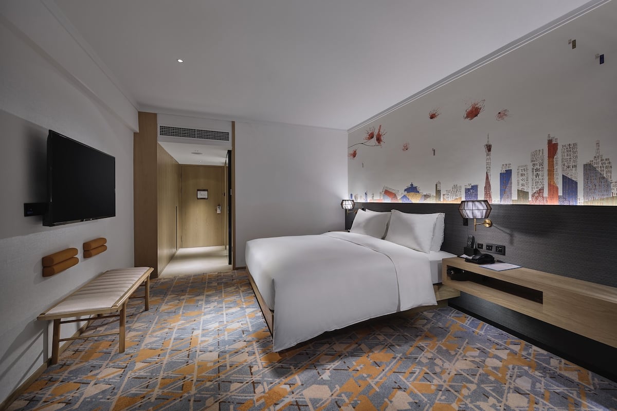 A hotel room with a bed, a TV, a bench, a desk, and a wall with a cityscape mural.