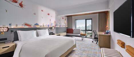 A hotel room with a large bed, a desk, a chair, a TV, and a cityscape mural on the wall.
