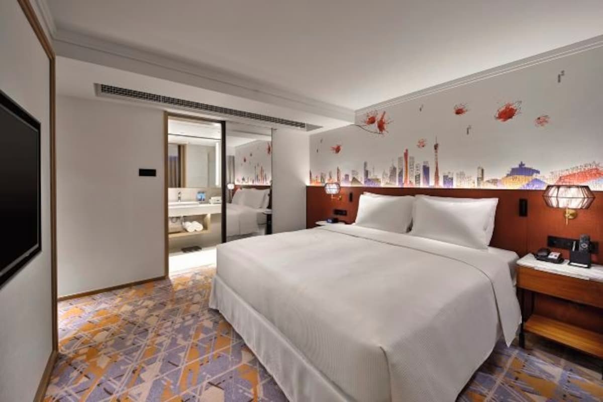 A hotel room with a large bed, a nightstand, a desk, and a mirror. There is a cityscape mural on the wall and a patterned floor.