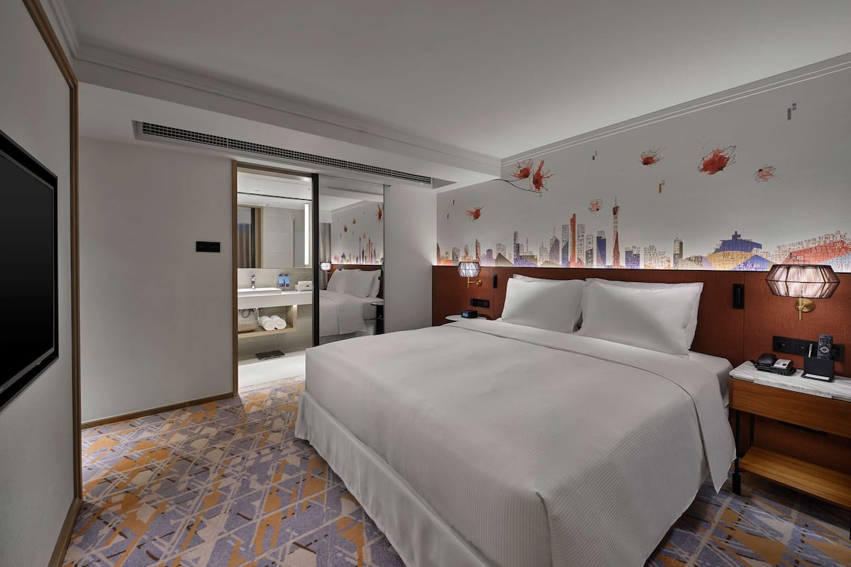 A hotel room with a large bed, a bedside table with a lamp, a wall-mounted TV, and a bathroom visible through an open door.