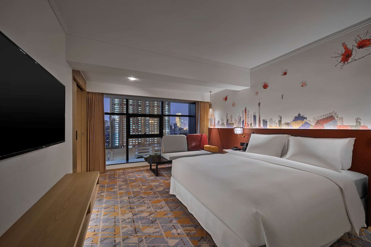 A hotel room with a large bed, a TV, a desk, and a view of a cityscape through large windows.