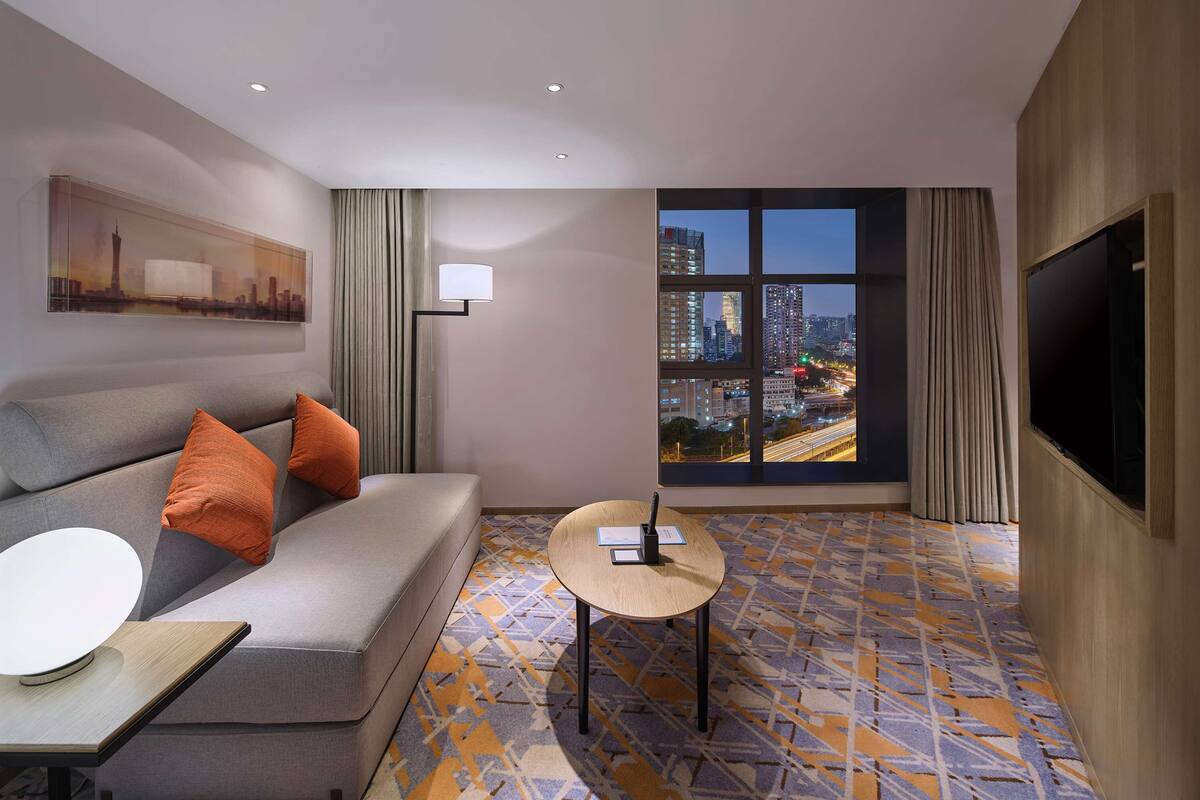 A hotel room with a sofa, a small table, a lamp, a painting, a large window with a city view, and a flat-screen TV.