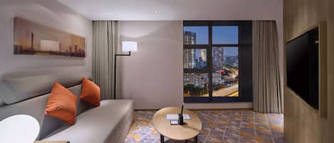 A hotel room with a sofa, a small table, a lamp, a painting, a large window with a city view, and a flat-screen TV.