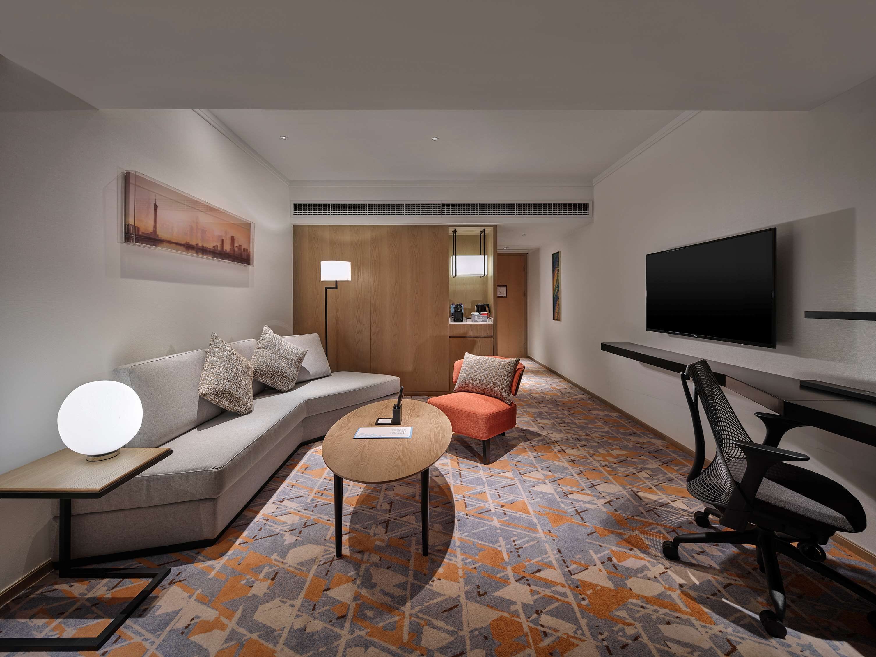Une chambre d’hôtel moderne, équipée d’un canapé, d’une petite table ronde, d’une chaise, d’un téléviseur et d’un tapis à motifs.