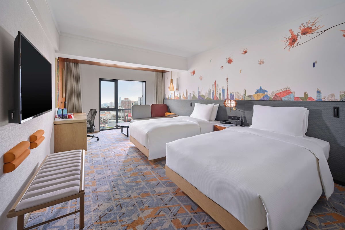 A hotel room with two beds, a desk, a chair, a TV, and a cityscape wall mural.