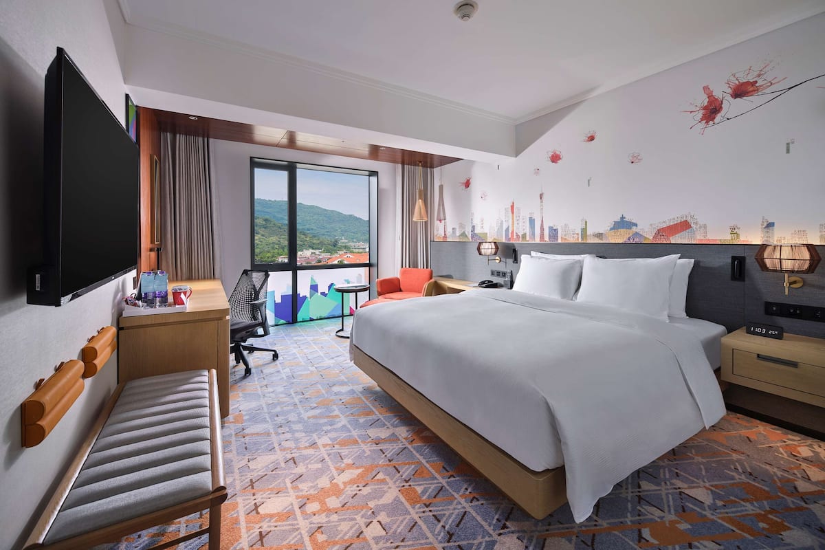 A hotel room with a large bed, a desk, a chair, a TV, and a balcony with a cityscape view.
