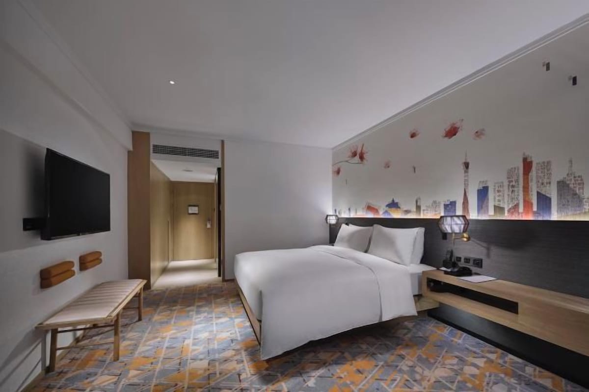 A hotel room with a bed, a bench, a TV, and a cityscape mural on the wall.
