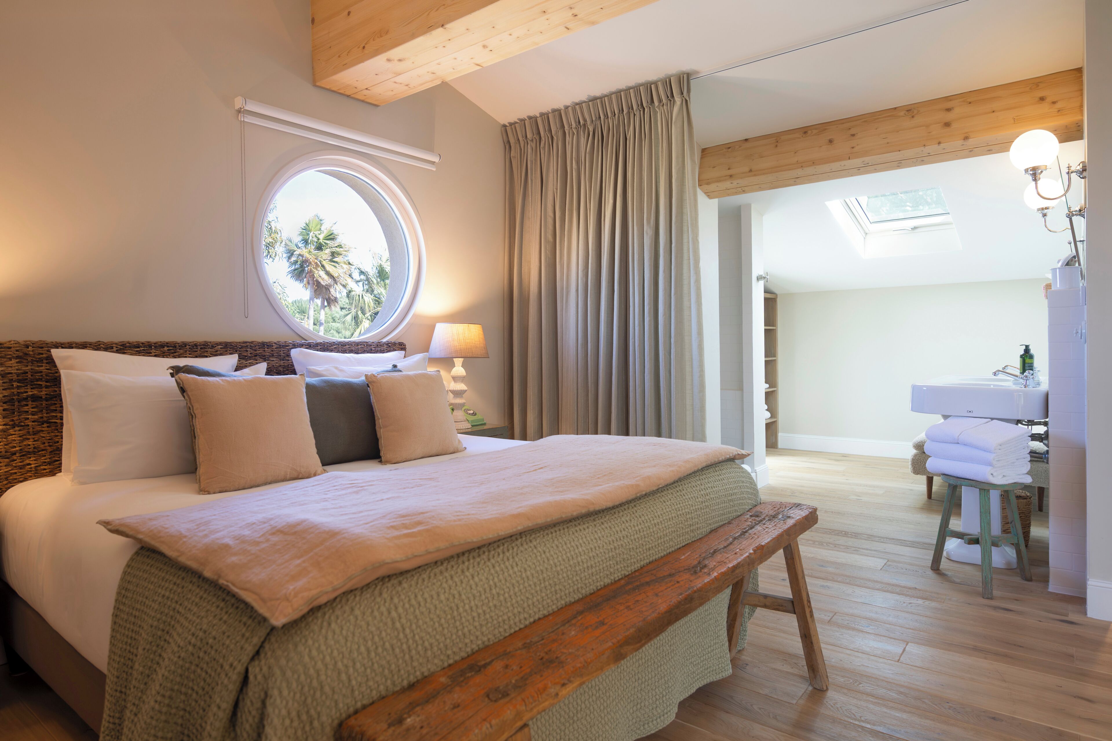 A bedroom with a large bed, a wooden bench, a round window, a bathroom with a sink and towels.