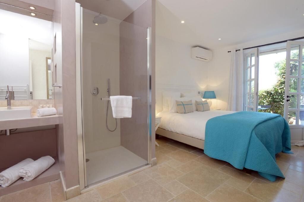 A modern hotel room with a glass-enclosed shower, a large bed with a blue bedspread, a bedside table with a lamp, and a view of greenery through the window.
