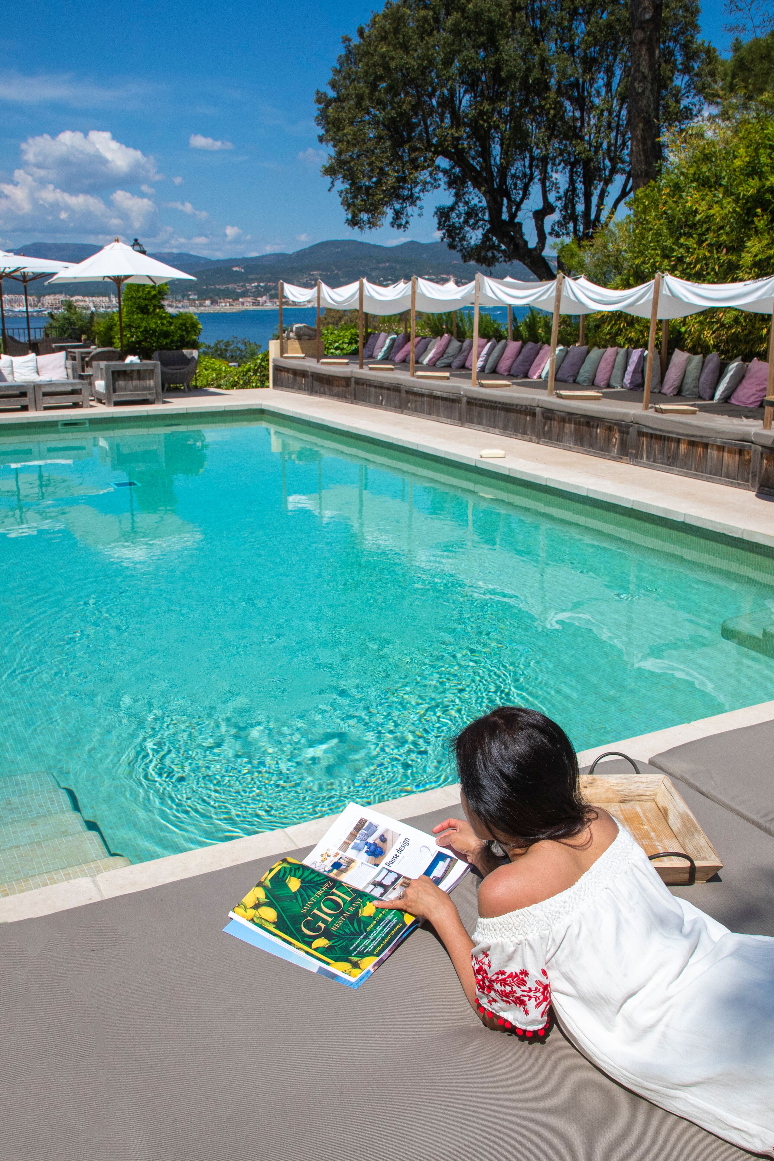 A poolside area with lounge chairs and umbrellas, a woman reading a book, and a view of the sea and hills in the background.