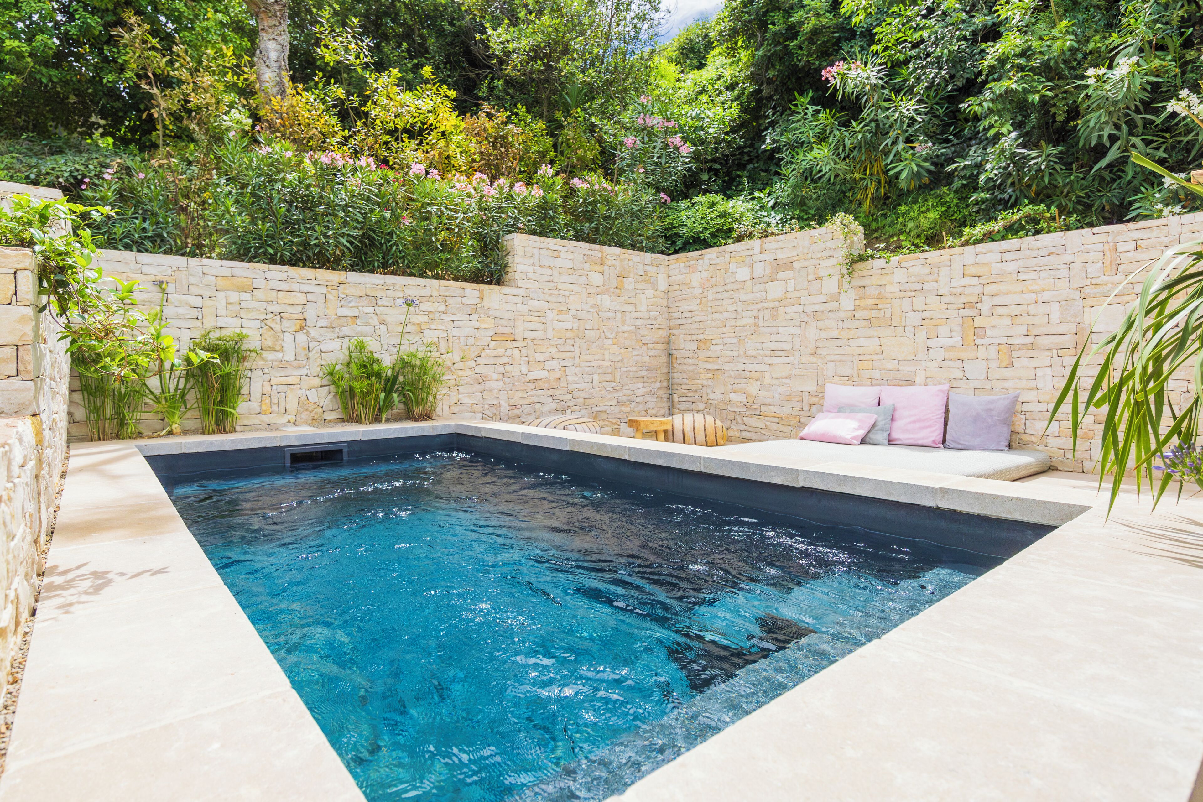A rectangular swimming pool with a stone wall surrounding it, a seating area with cushions, and lush greenery in the background.