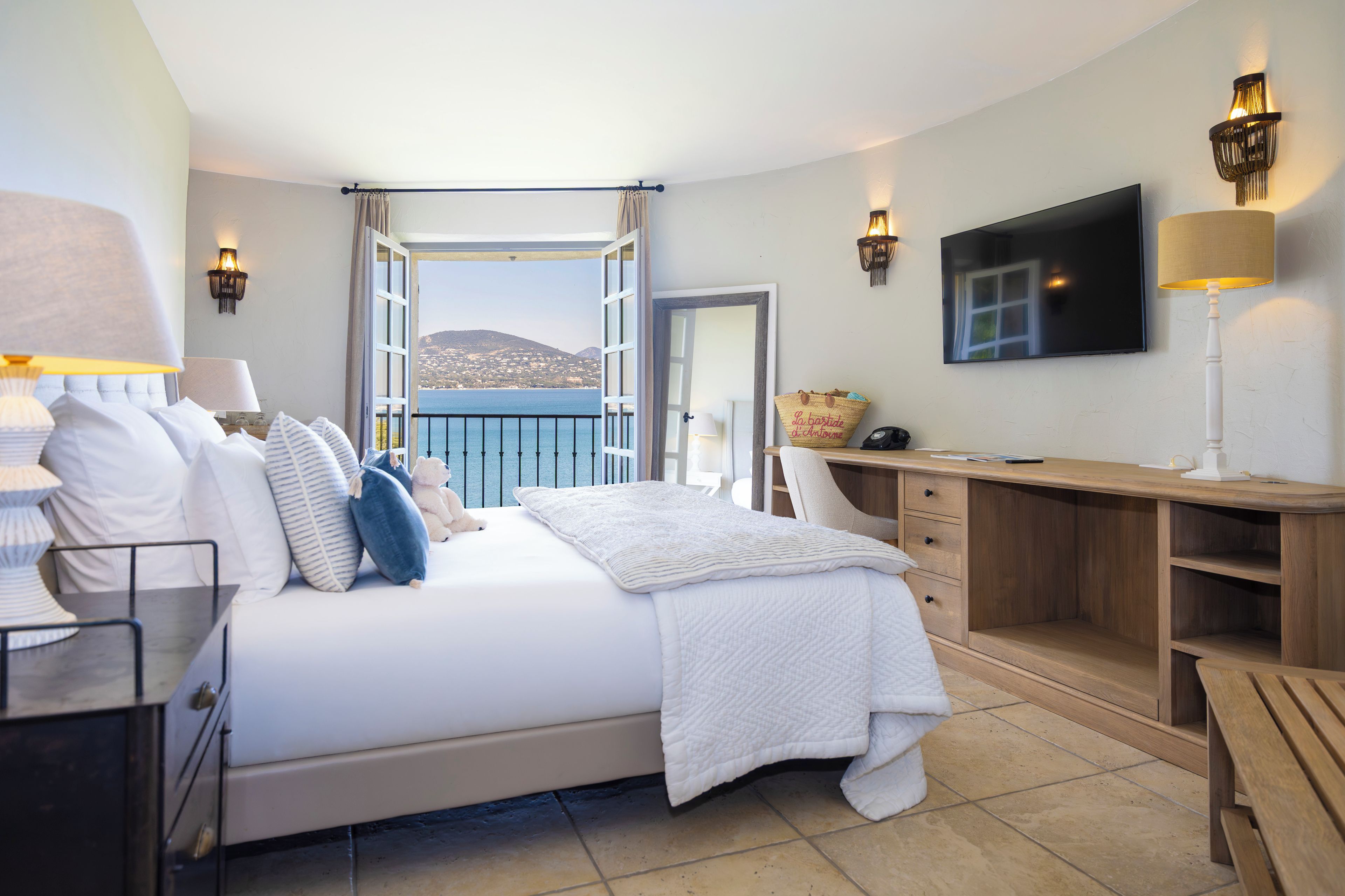 A bedroom with a bed, bedside tables, a desk, a TV, and a view of the sea.