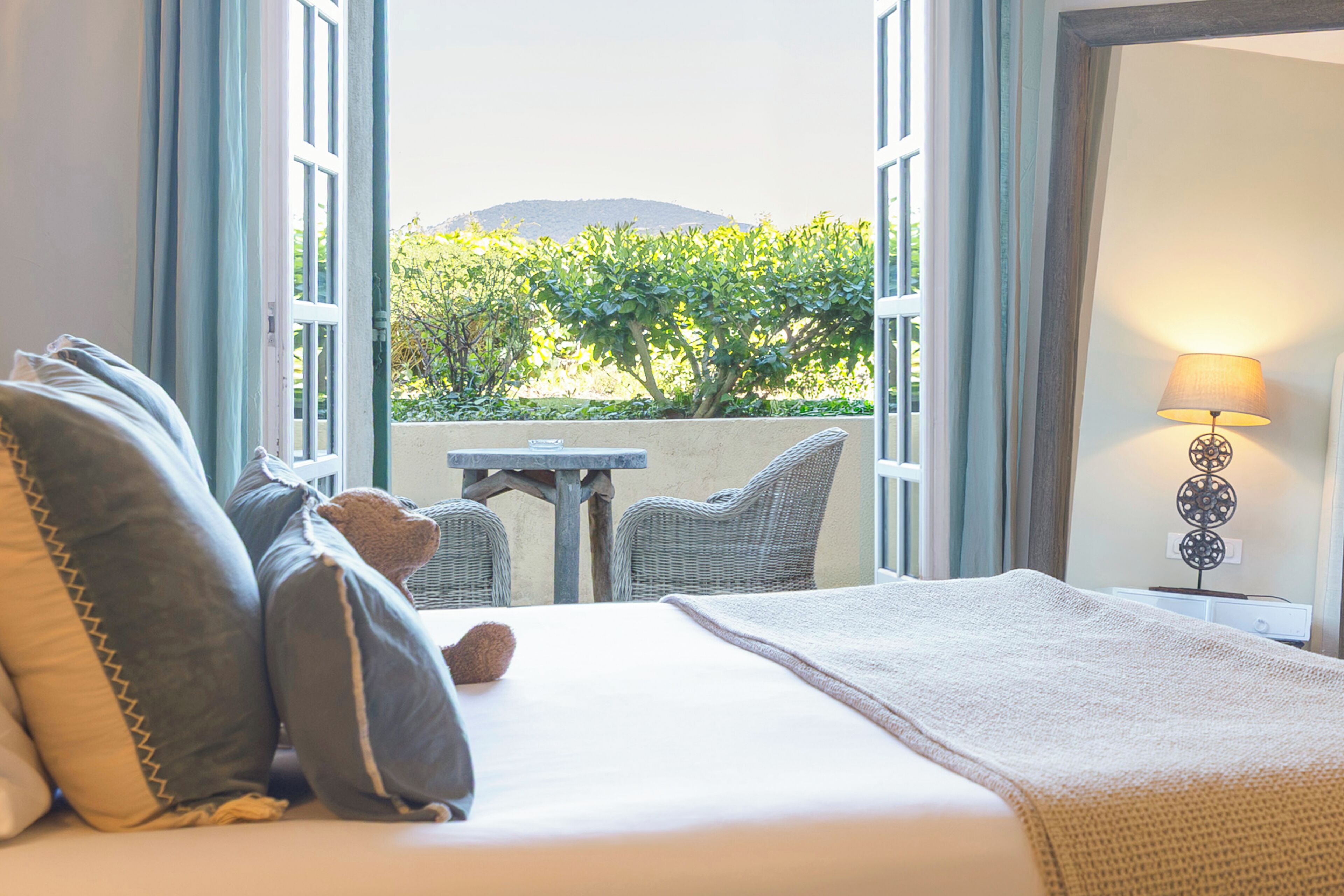 Ein Bett mit Kissen, ein Nachttisch, eine Lampe und Blick ins Grüne durch offene Fenster.