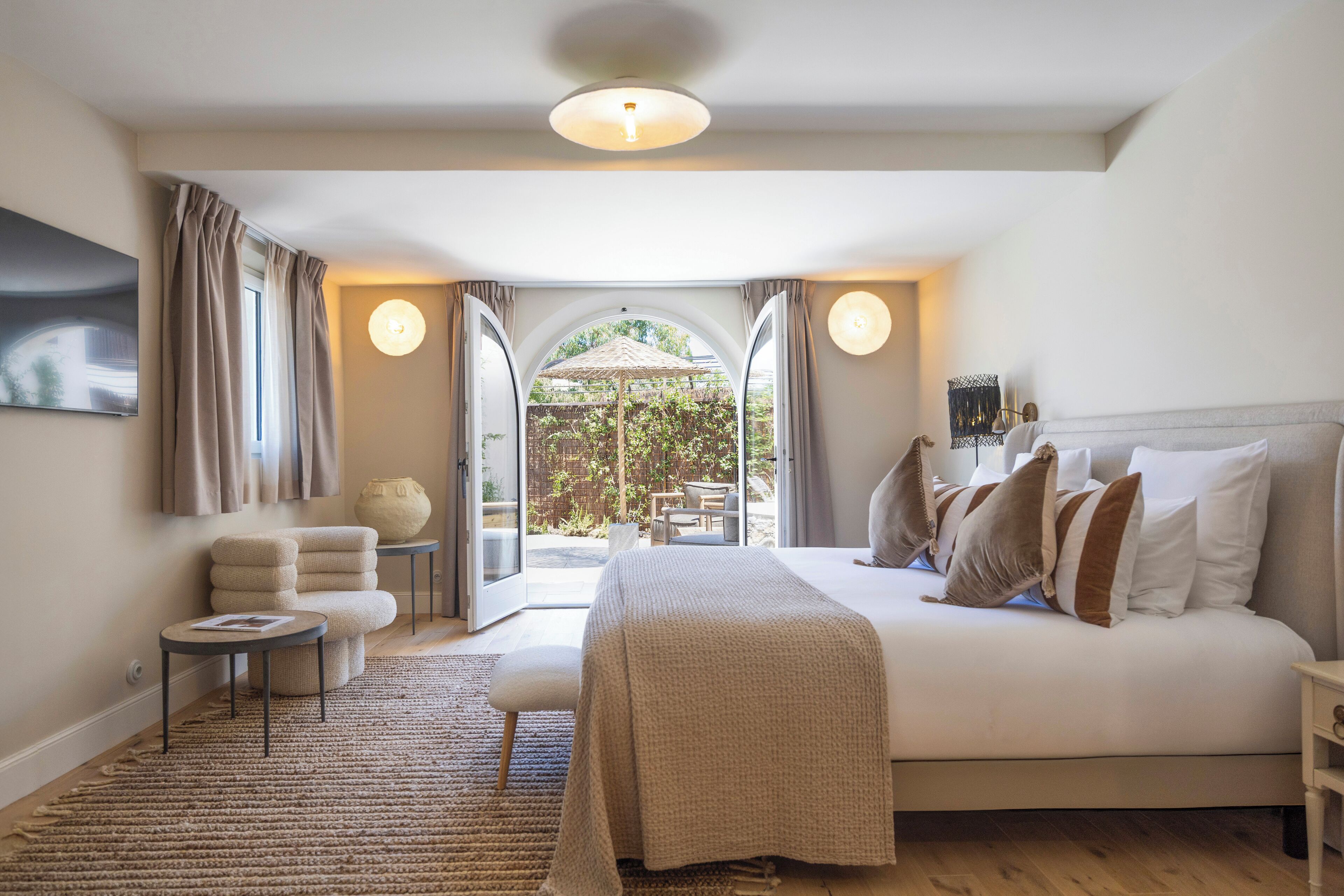 Een slaapkamer met een groot bed, een klein rond tafeltje, een stoel en uitzicht door een open deur op een buitenruimte met een tafel en stoelen.