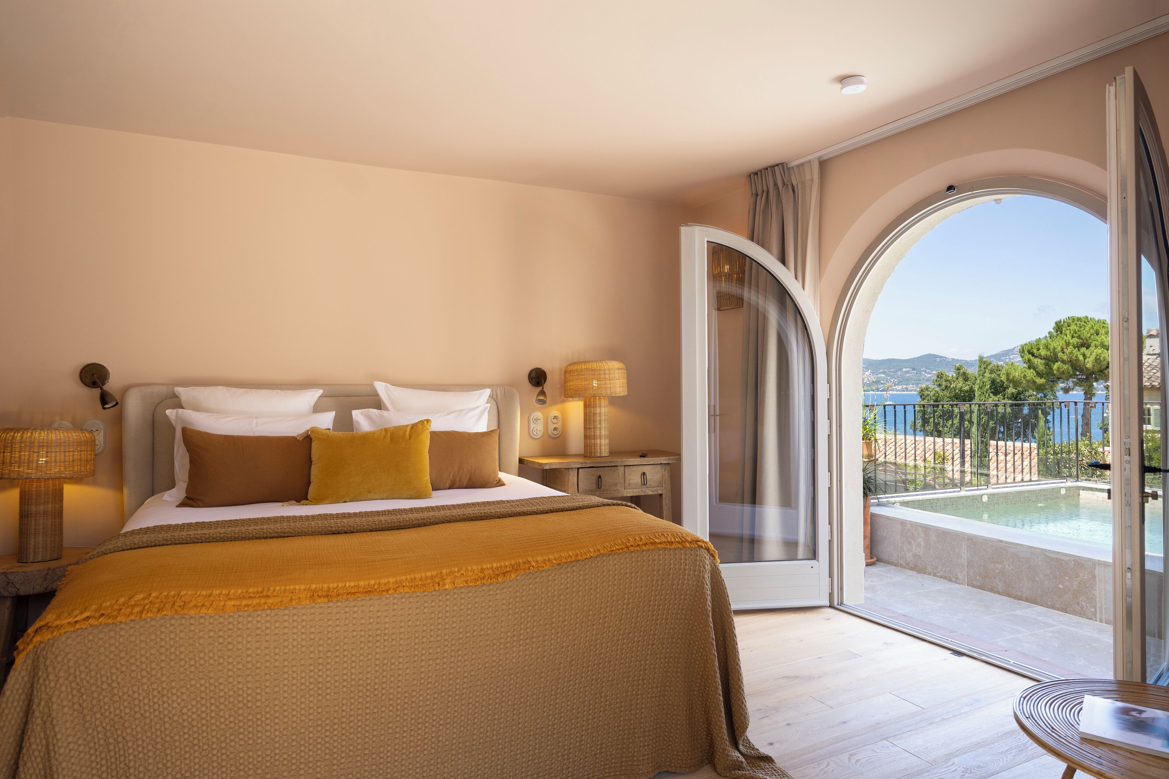 A bedroom with a large bed, a nightstand, a lamp, and a view of a swimming pool through an open door.