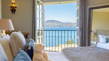 A hotel room with a bed, a sofa, a teddy bear, and a balcony with a view of the sea and a distant town.