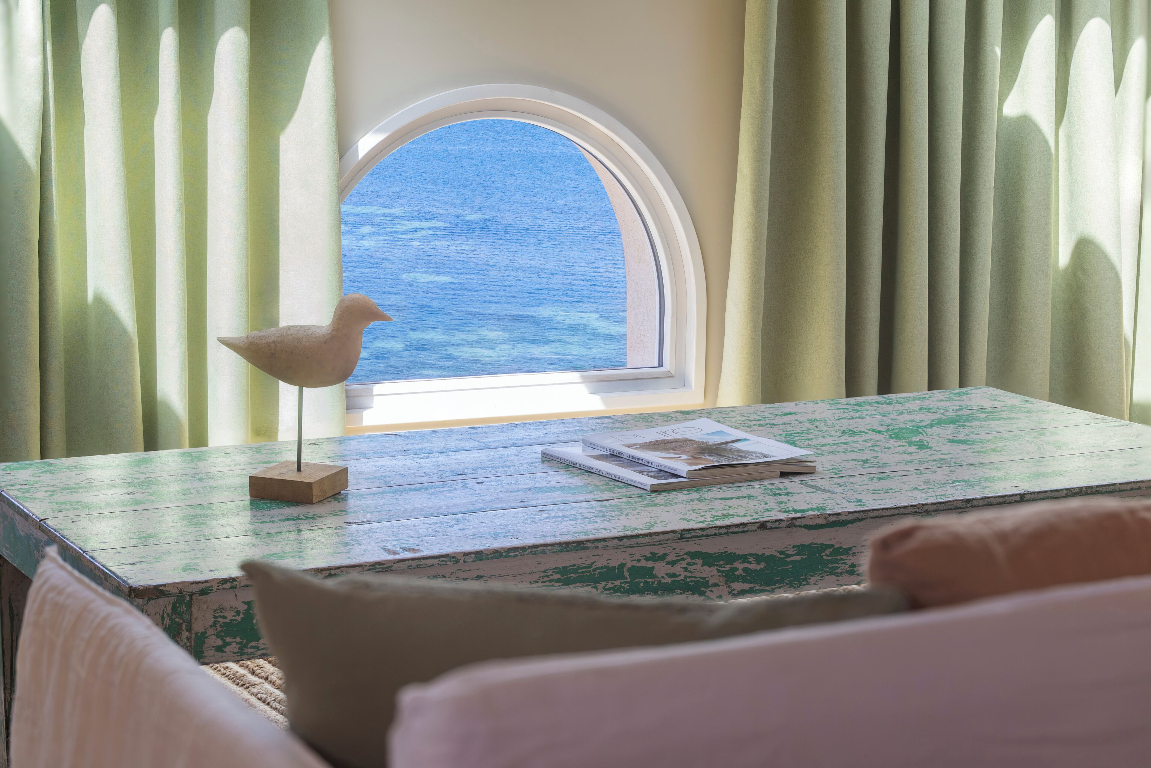 A room with a view of the sea through a window, a table with a bird sculpture and a book, and a bed with pillows.