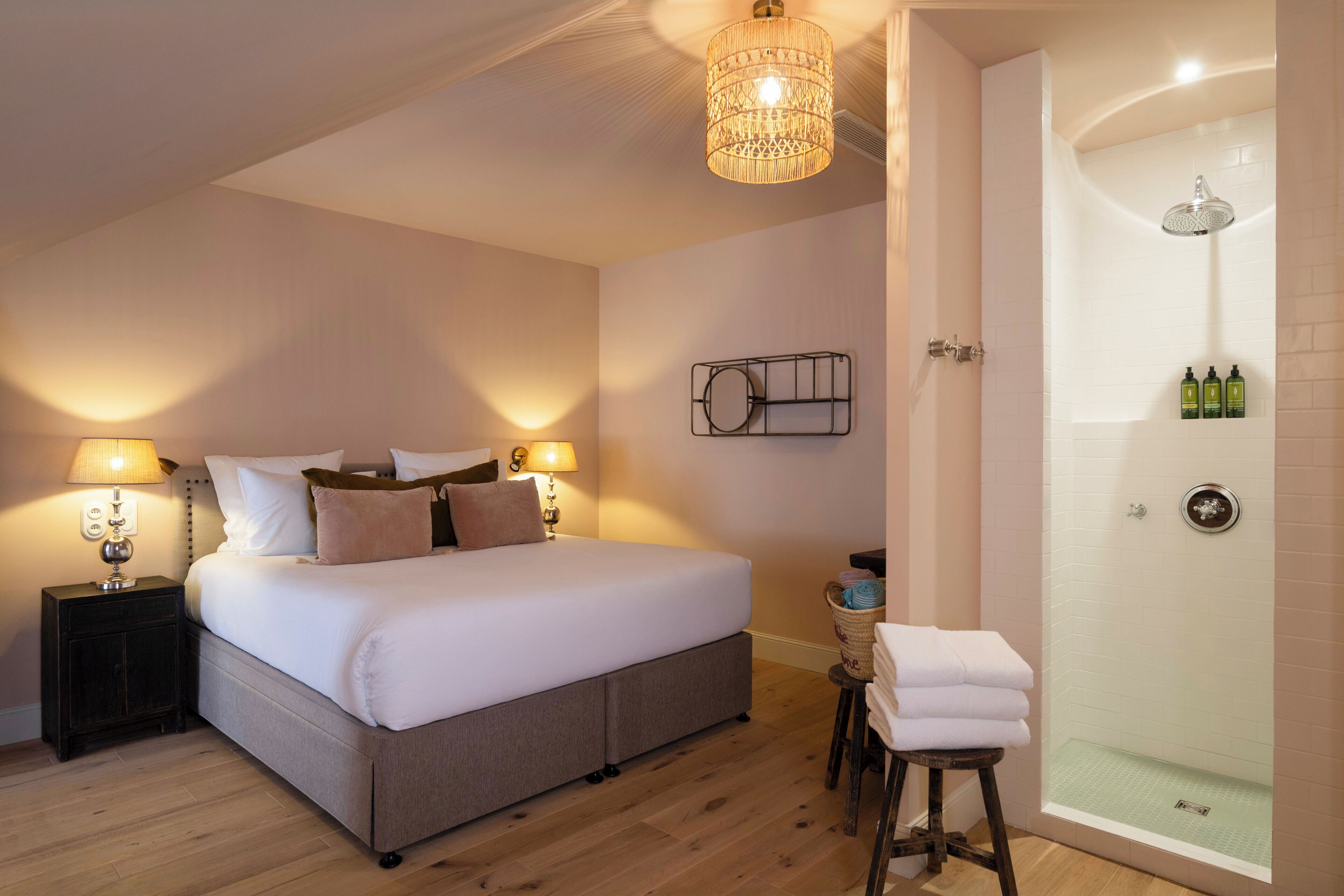A bedroom with a large bed, bedside lamps, a nightstand, a small table with a basket, and a shower area with a glass door.