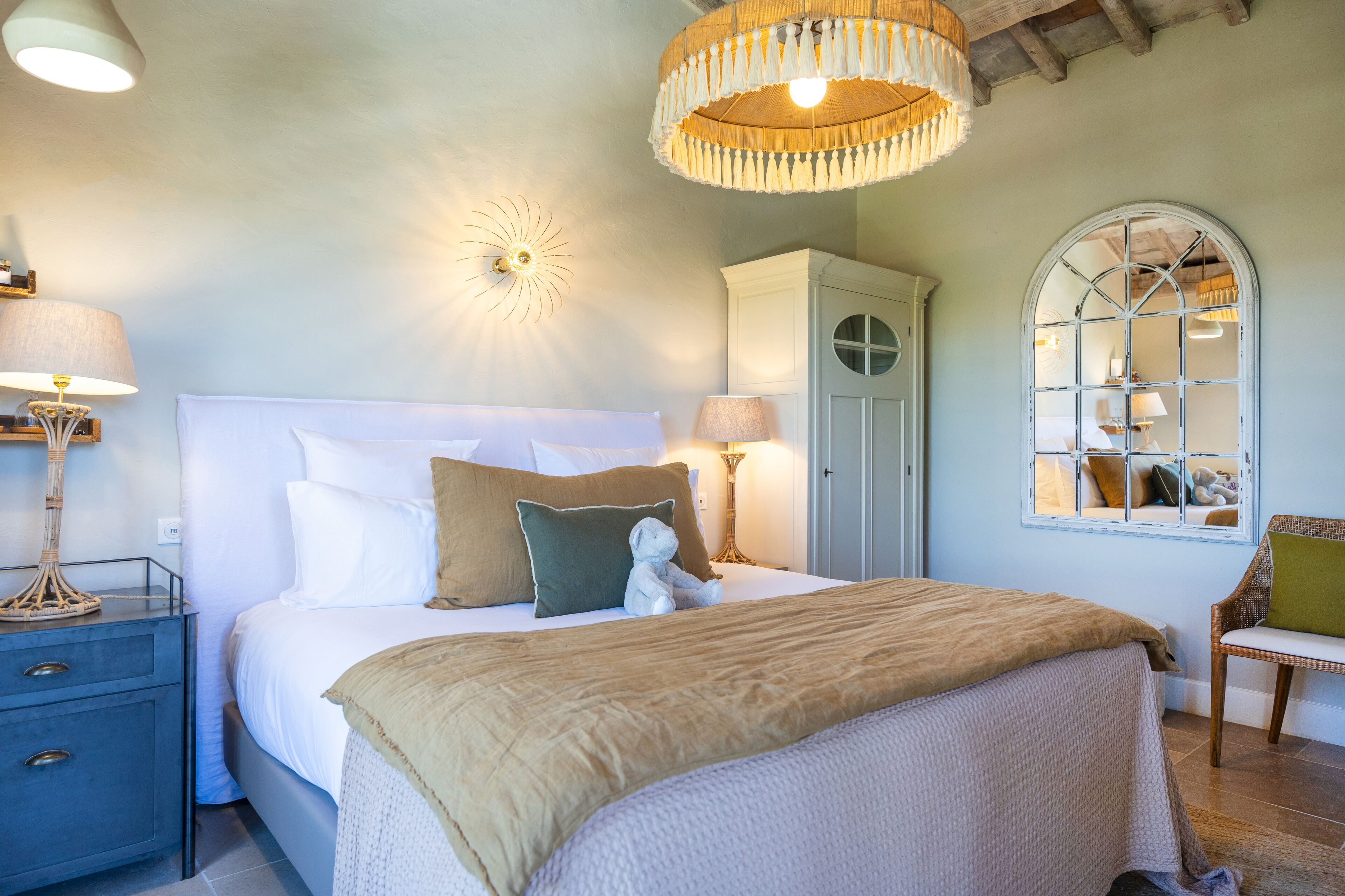 A bedroom with a large bed, bedside lamps, a decorative wall light, a window with a view, and a wicker chair.