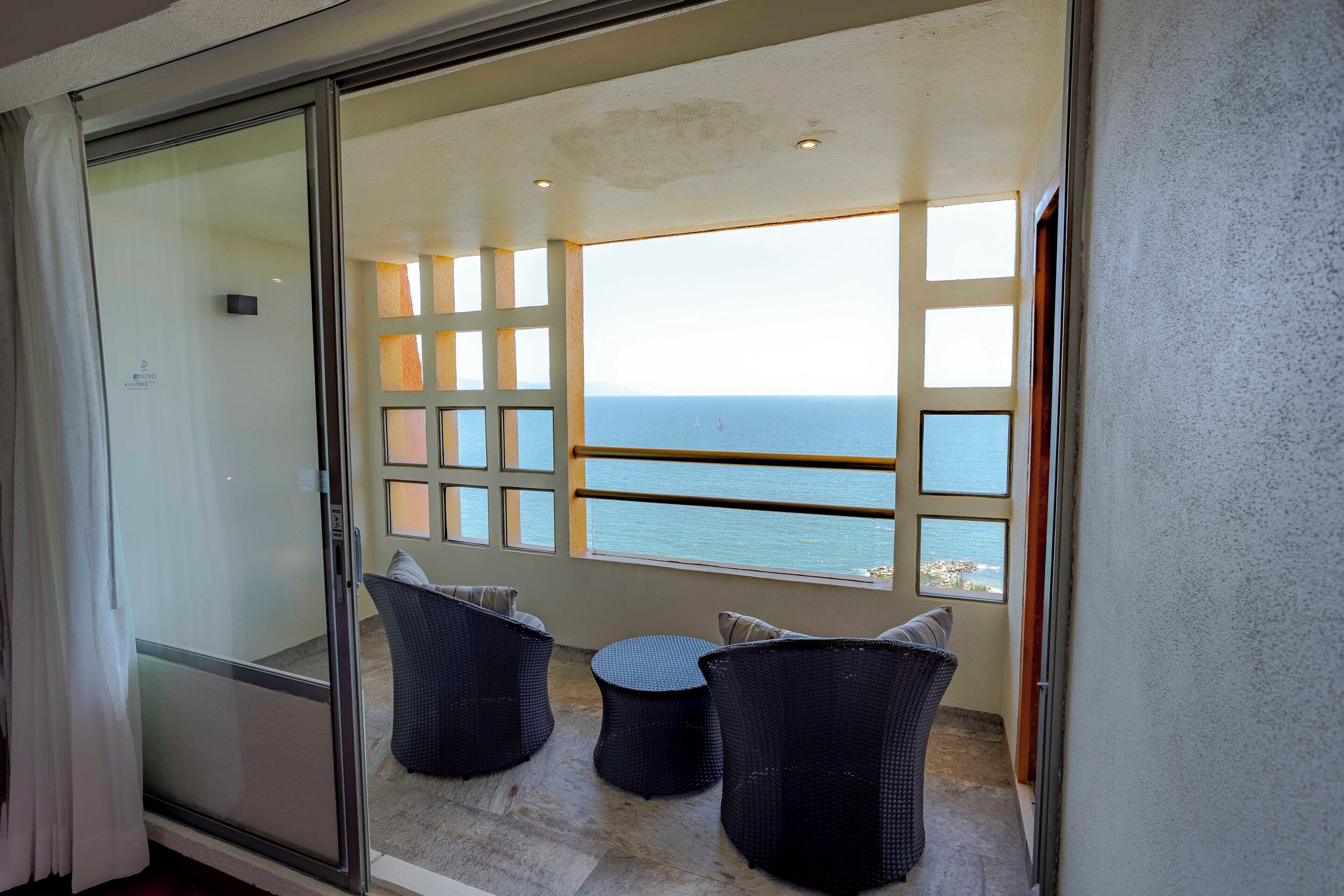 A balcony with a view of the sea, featuring a glass door, two wicker chairs, and a railing.