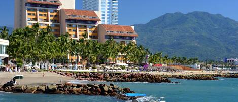 Un hotel frente al mar con varios pisos, una playa al fondo y un litoral rocoso.