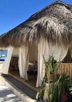 Uma cabana de praia com telhado de palha, cortinas brancas e um banco de madeira.