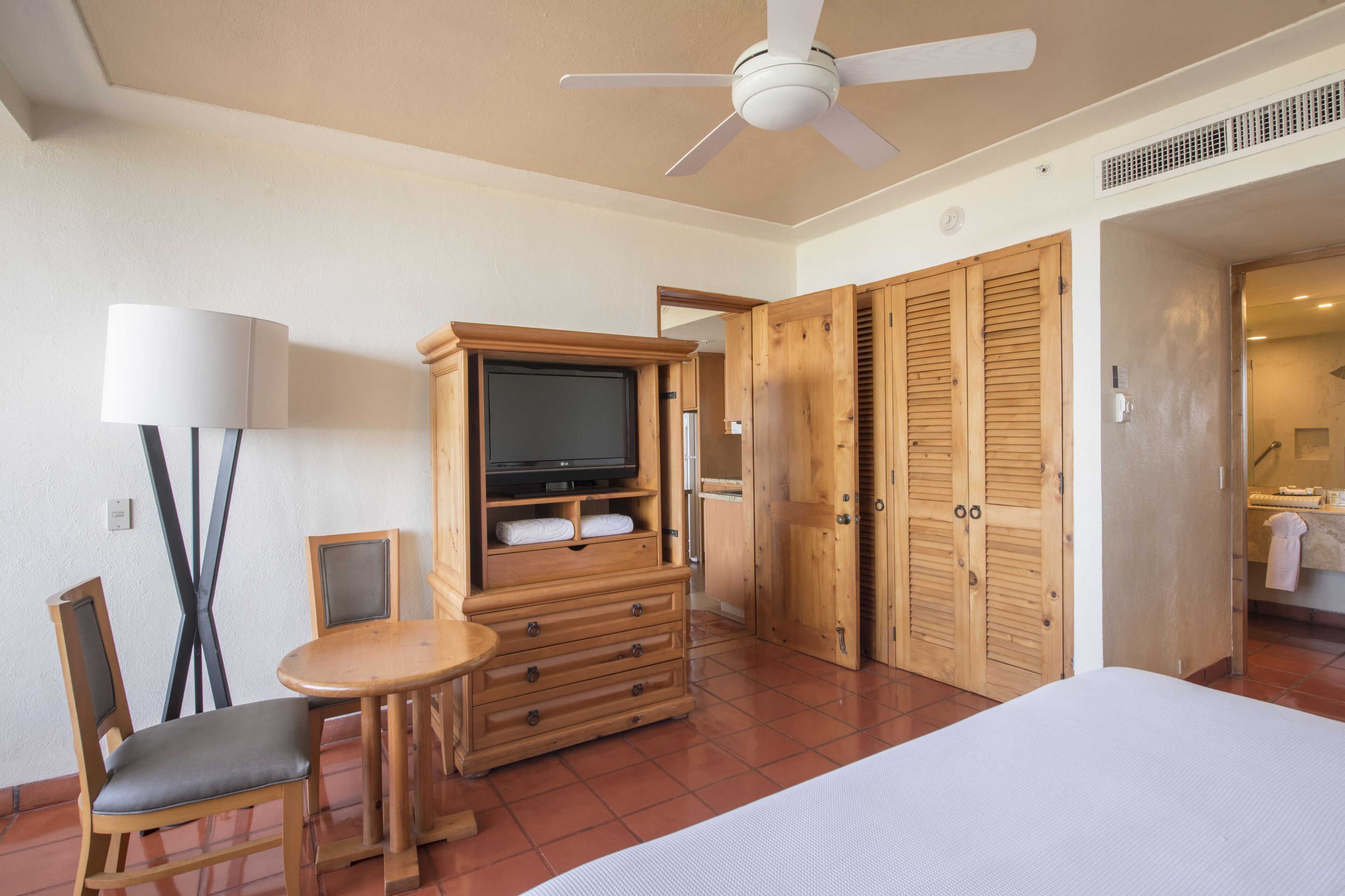 A hotel room with a bed, a TV, wooden furniture, a ceiling fan, and a bathroom visible through an open door.