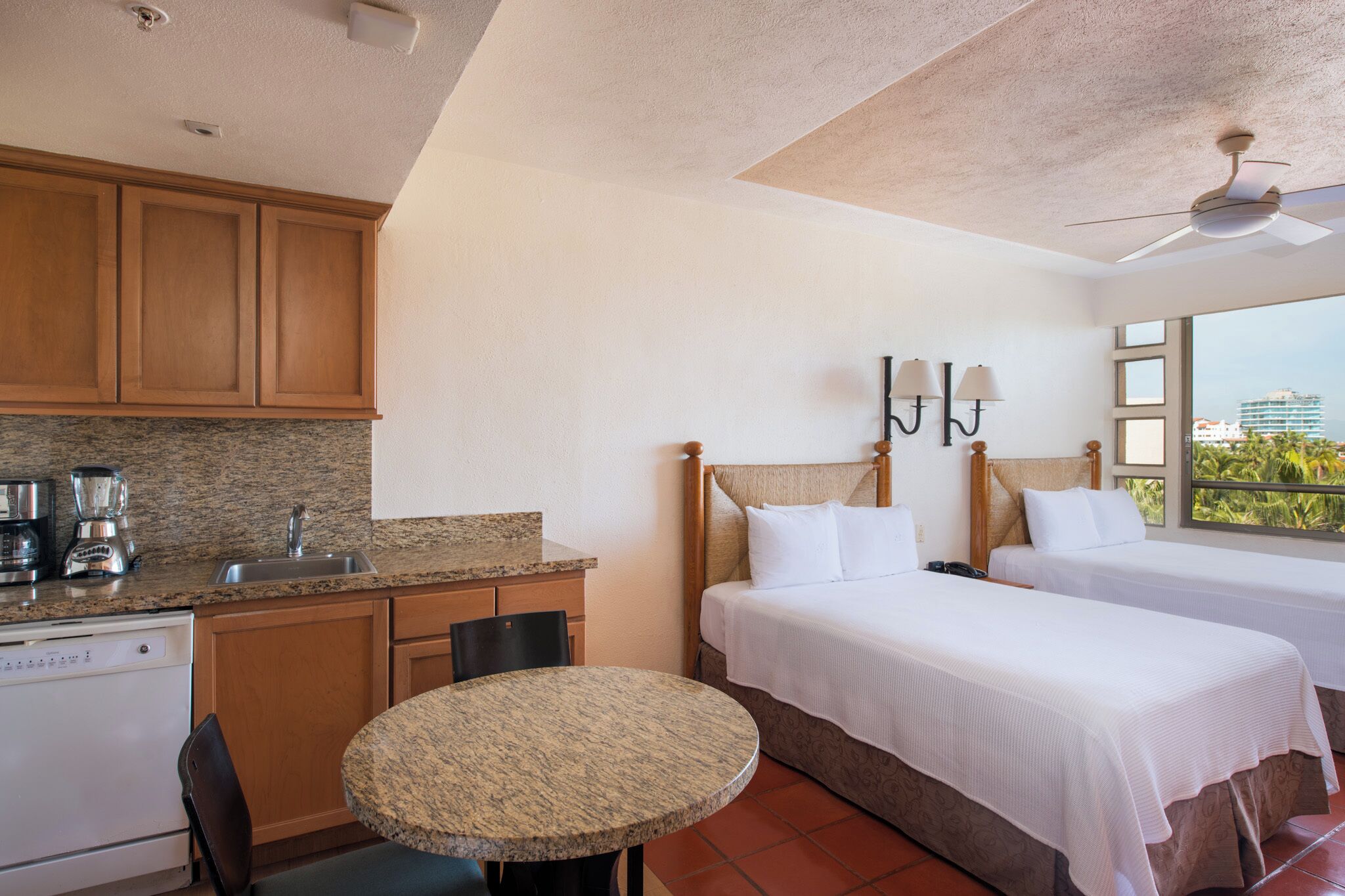 Une chambre d’hôtel avec deux lits, une petite cuisine équipée d’un lave-vaisselle, une table à manger et une grande fenêtre donnant sur un paysage agréable.