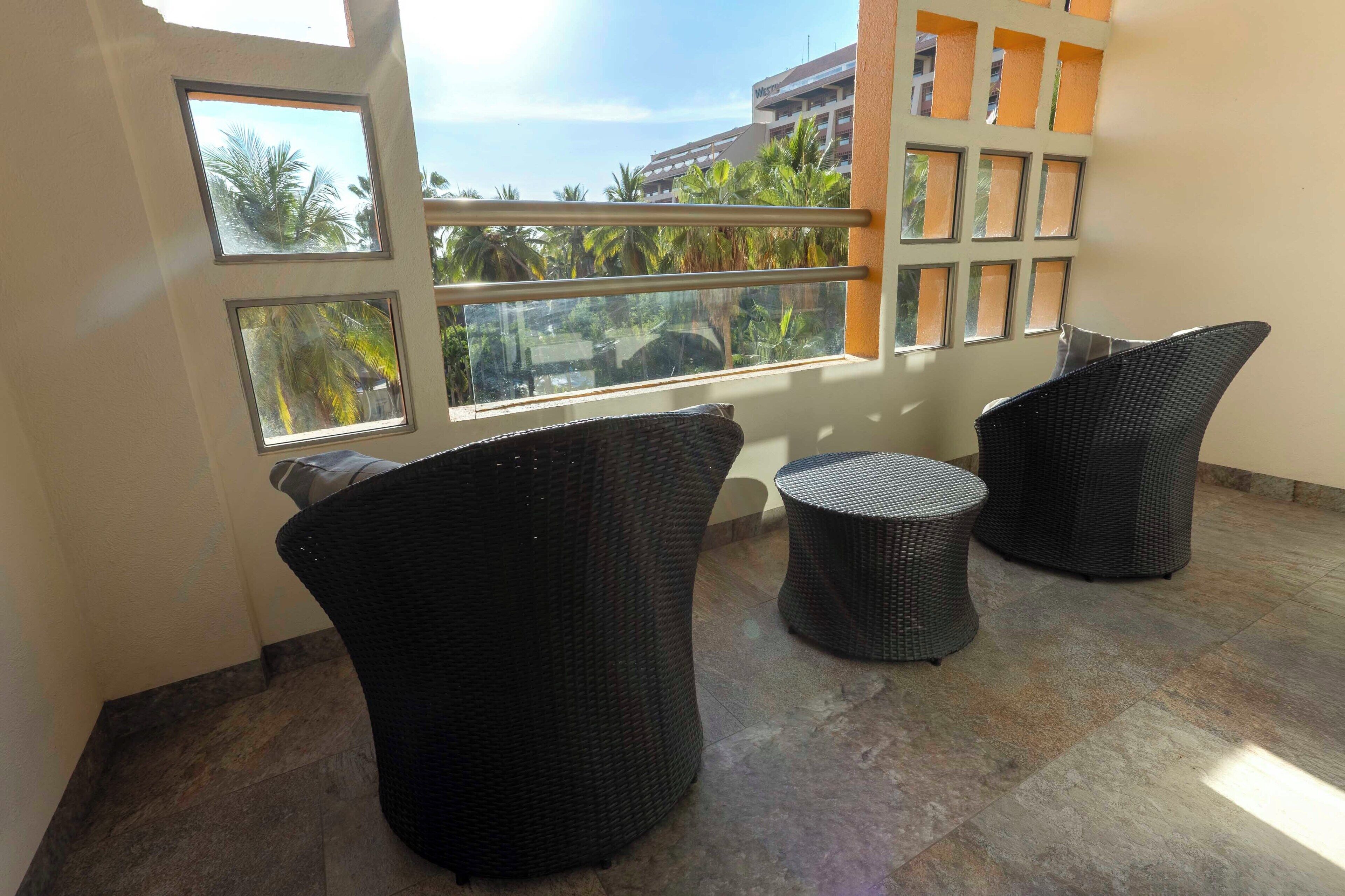 A balcony with two wicker chairs and a small round table, overlooking a building with palm trees.