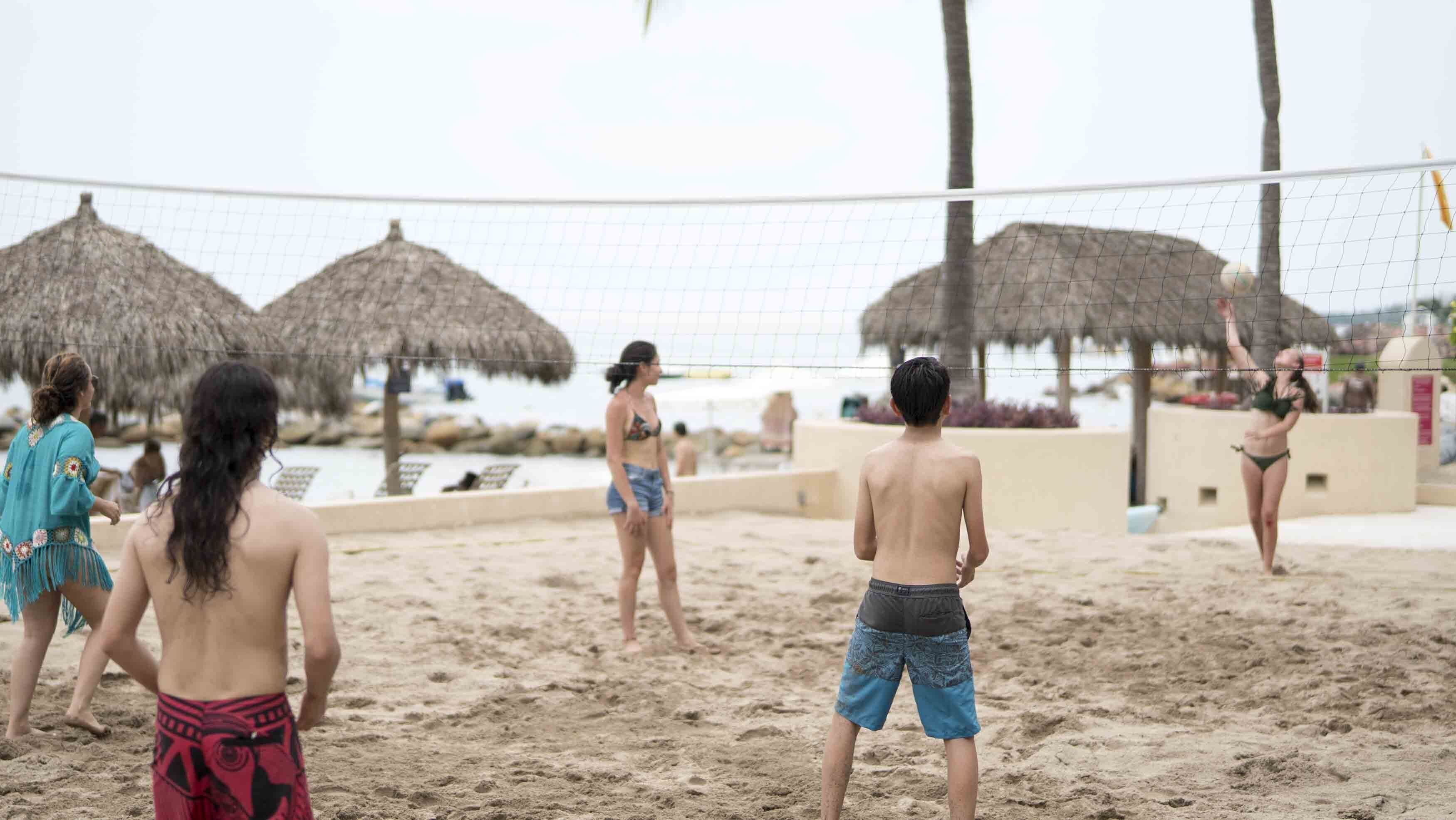 Partita di beach volley con giocatori in costume da bagno, una rete da pallavolo e ombrelloni di paglia sullo sfondo.