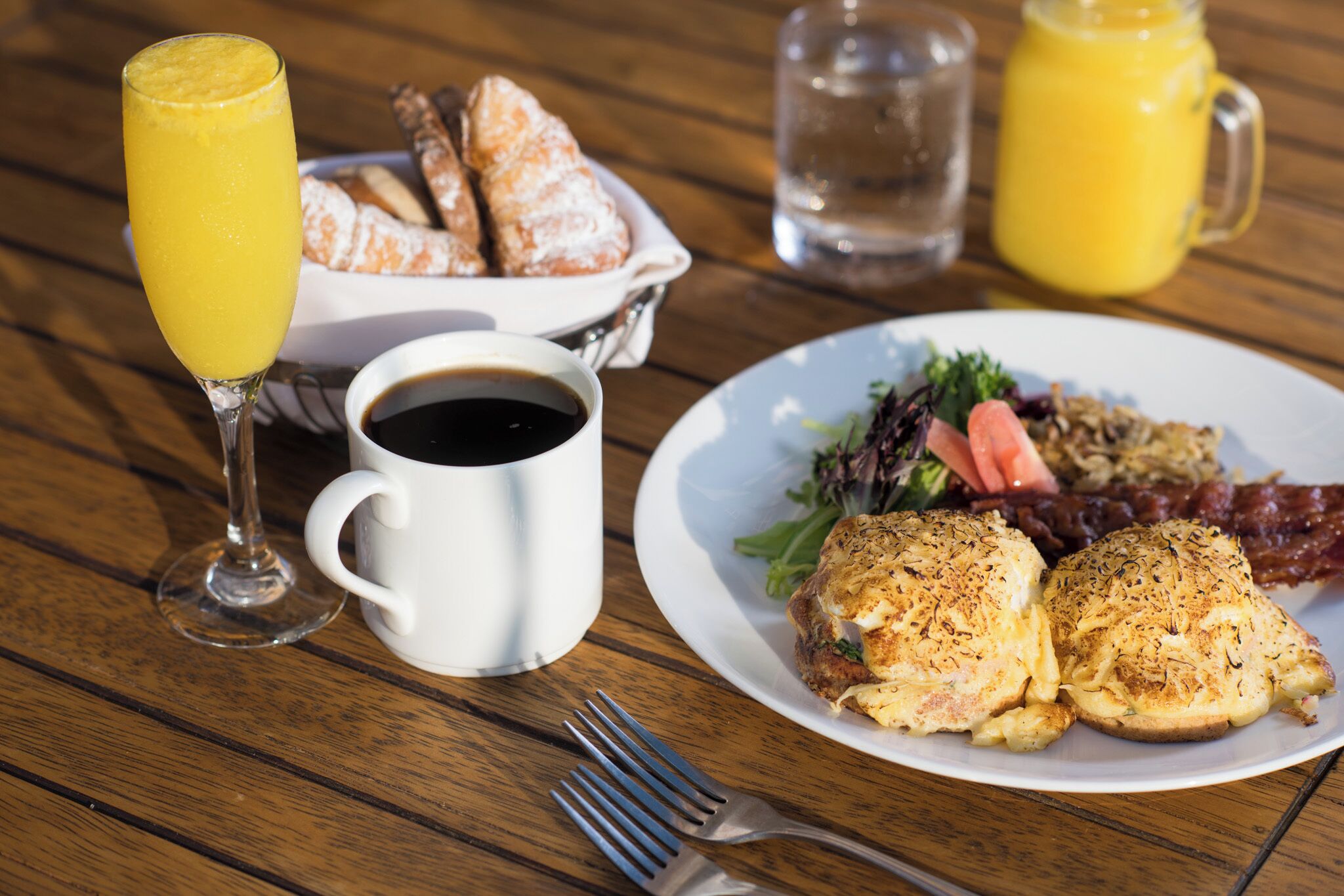 A breakfast setting with croissants, a plate of eggs, a glass of orange juice, a cup of coffee, and a pitcher of water on a wooden table.