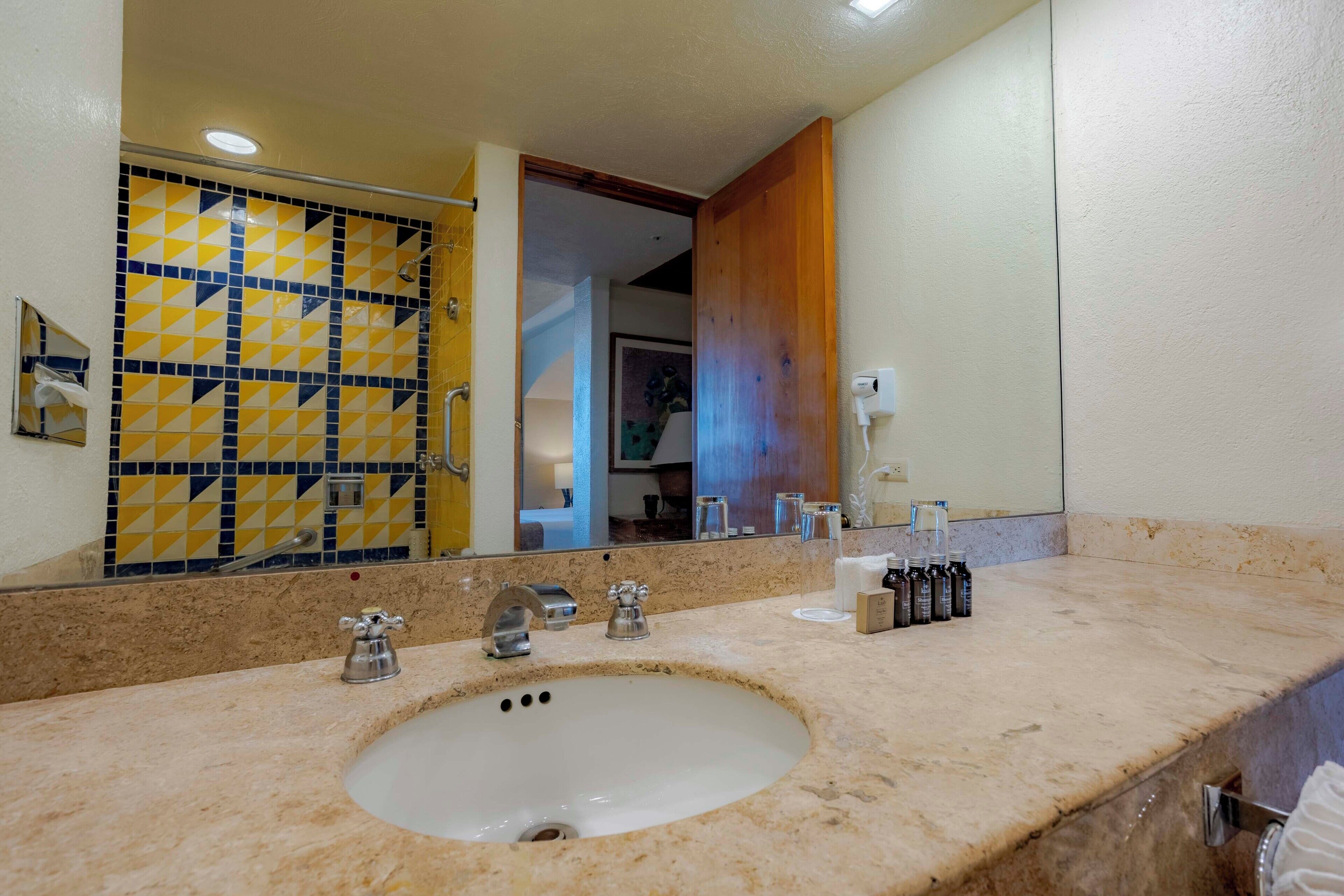 A bathroom with a sink, three faucets, a mirror, and a shower area with yellow and blue tiles.