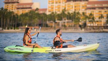 Dos personas haciendo kayak en aguas tranquilas, con un hotel y palmeras al fondo.