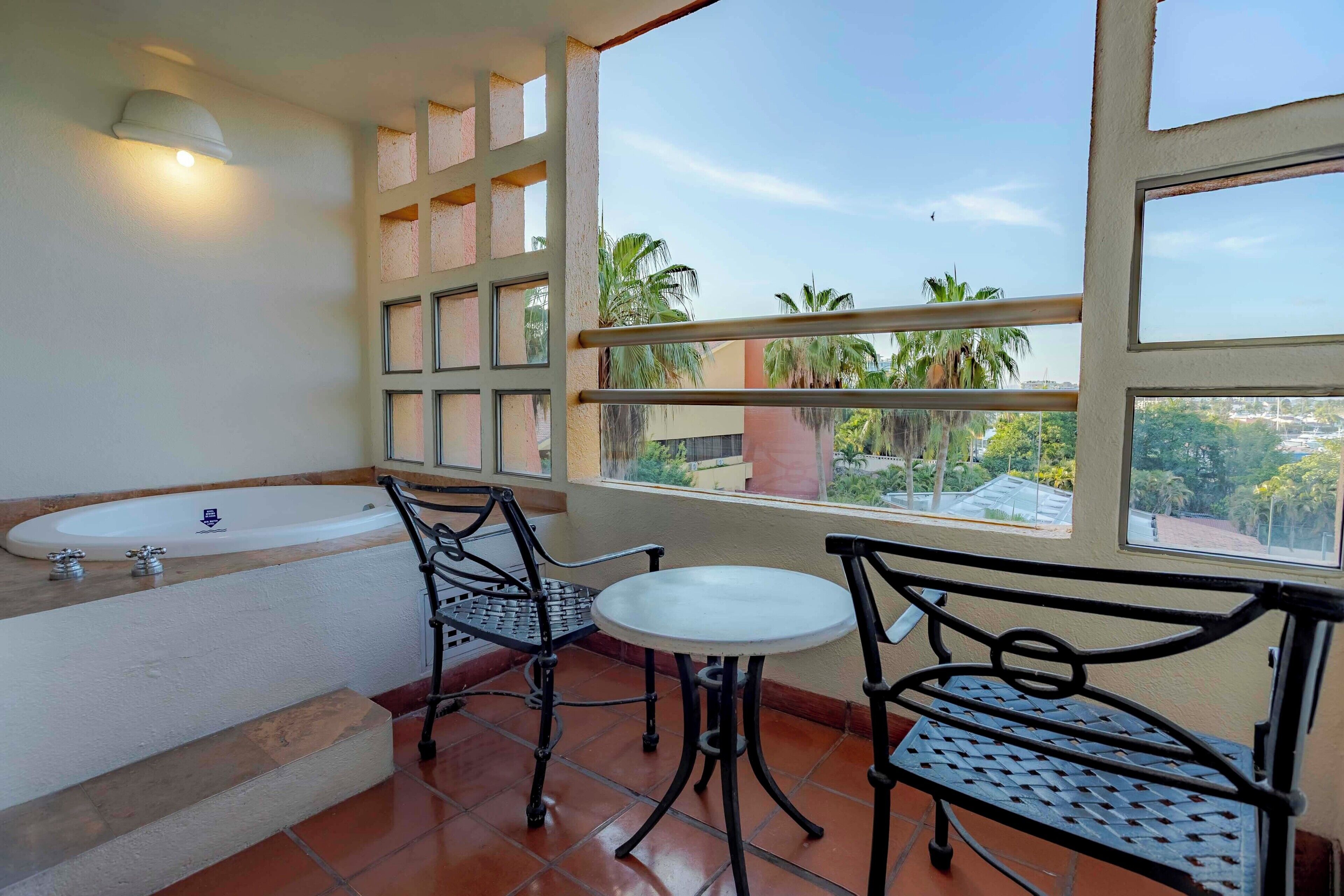 A balcony with a view of palm trees and buildings, a small table, two chairs, a sink, and a window with multiple panes.