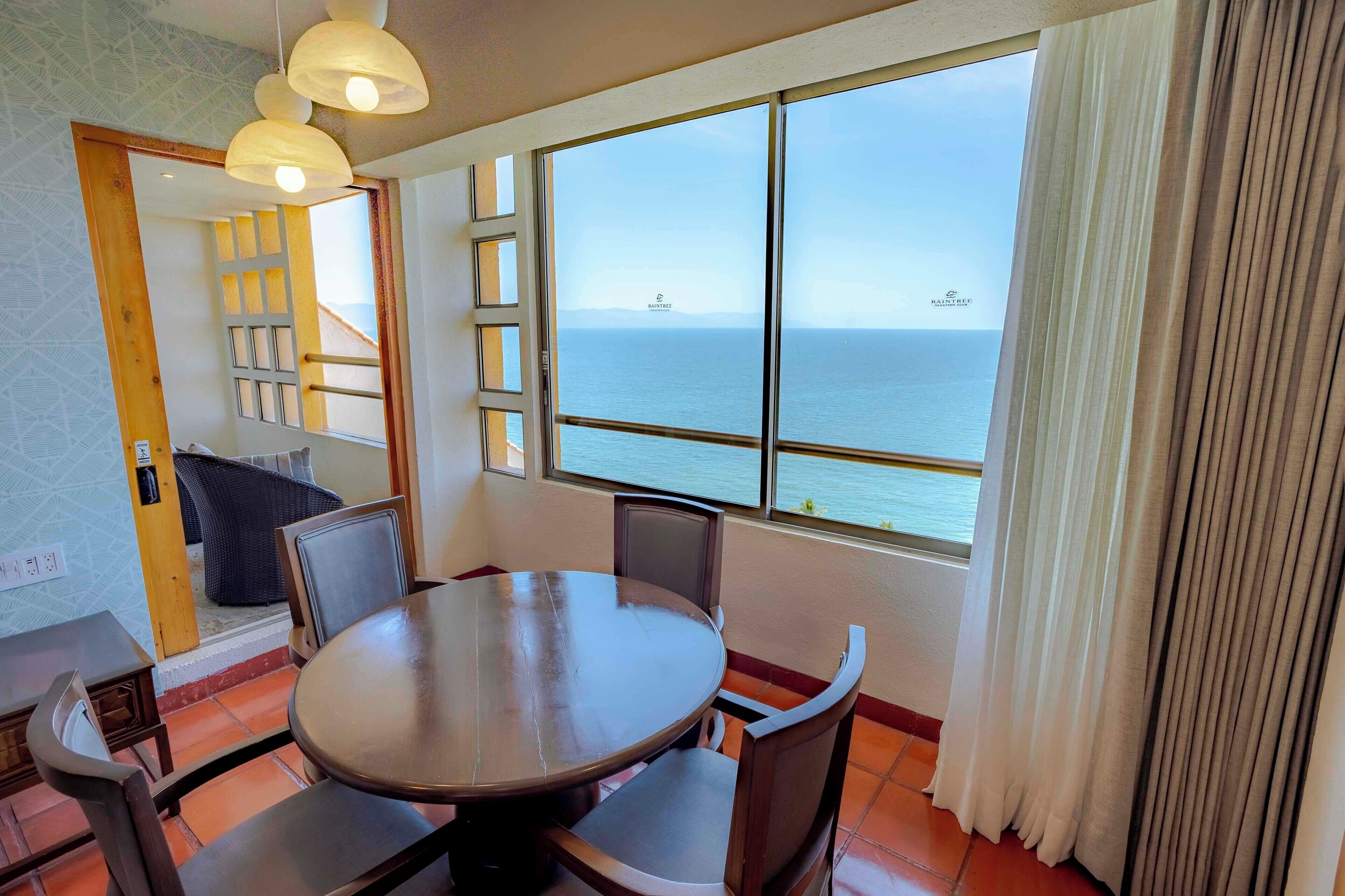 A room with a round table, chairs, a view of the sea, and a window with curtains.