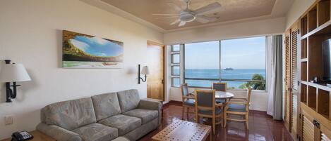 Un soggiorno con divano, tavolino e zona pranzo con sedie e tavolo. È presente una ventola a soffitto, un quadro appeso al muro e vista sull'oceano attraverso ampie finestre.