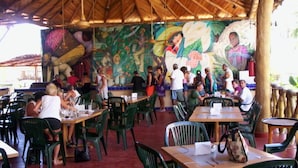 An outdoor restaurant with wooden tables and chairs, a thatched roof, and a mural on the wall. - El Tropicano (Acapulco)