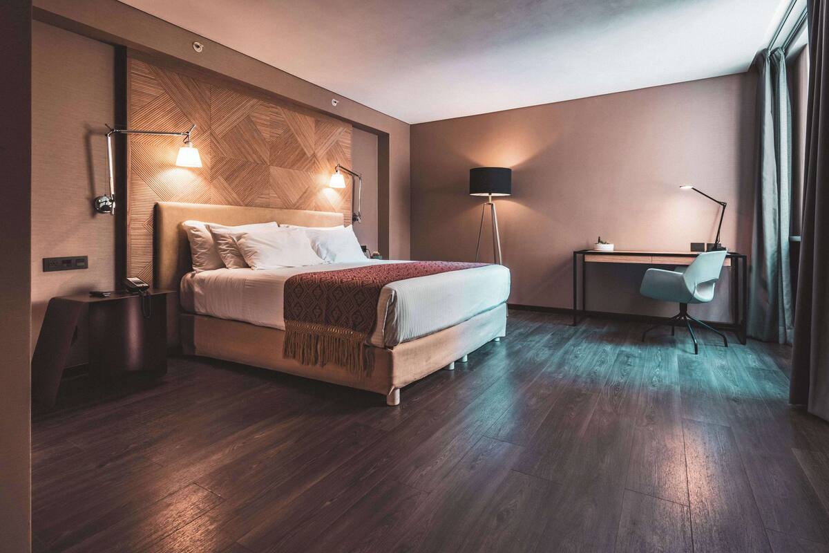 Een moderne hotelkamer met een groot bed, nachtlampen, een bureau met stoel en uitzicht op het omliggende landschap.