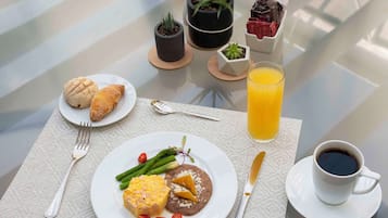 A breakfast setting with croissants, a plate of eggs, beans, asparagus, and a glass of orange juice on a table.