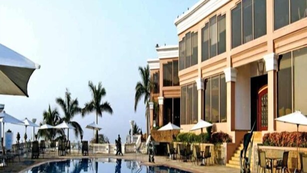 A swimming pool with lounge chairs and umbrellas, surrounded by palm trees and a building with large windows. - Palms Hotel (Mumbai)