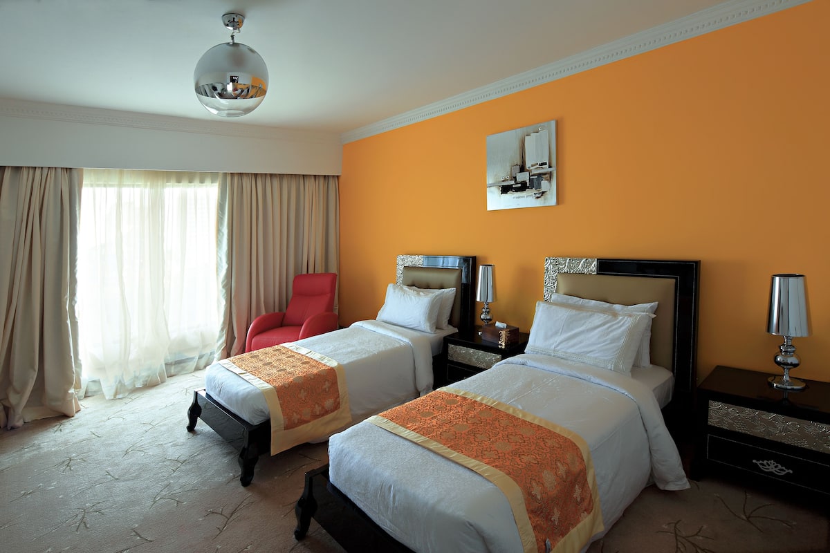 Habitación de hotel con dos camas, una silla roja y un ventanal con cortinas.